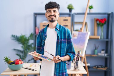 Young hispanic man artist holding paintbrush and palette at art studio
