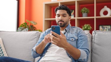Evdeki koltukta oturan Afro-Amerikan adam akıllı telefon kullanıyor.