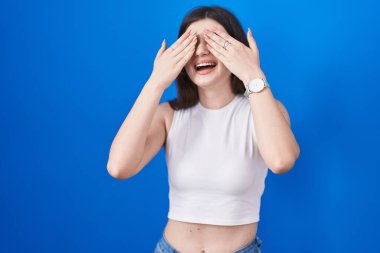 Young caucasian woman standing over blue background covering eyes with hands smiling cheerful and funny. blind concept. 
