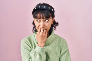 Young beautiful woman standing over pink background looking stressed and nervous with hands on mouth biting nails. anxiety problem. 