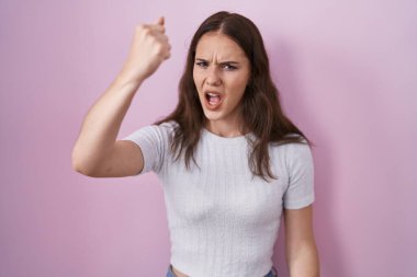 Genç İspanyol kız pembe arka planda kızgın ve kızgın duruyor öfkeyle bağırırken yumruğunu sallıyor ve öfkeleniyor. öfke ve agresif konsept. 