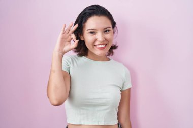 Pembe arka planda duran Latin kökenli genç kadın gülümsüyor ve el ve parmaklarıyla 