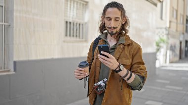 Genç İspanyol turist sokakta elinde kahve tutan akıllı telefon kullanıyor.