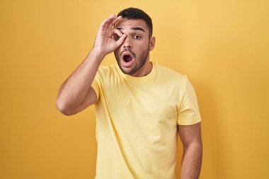 Young hispanic man standing over yellow background doing ok gesture shocked with surprised face, eye looking through fingers. unbelieving expression. 