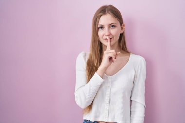 Young caucasian woman standing over pink background asking to be quiet with finger on lips. silence and secret concept. 