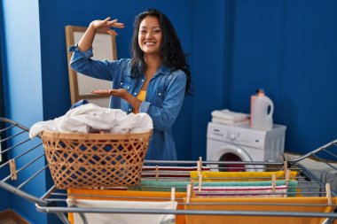 Genç Asyalı kadın çamaşır ipine elbise asıyor. Elleriyle işaret ediyor. Büyük ve büyük beden işareti, ölçü sembolü. Kameraya bakıp gülümsüyor. ölçüm kavramı. 