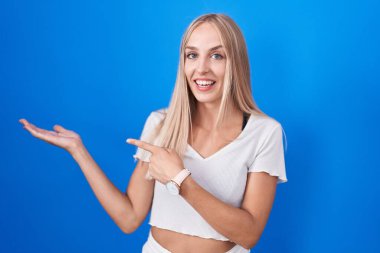 Mavi arka plan üzerinde duran genç beyaz kadın hayret içinde kameraya bakıp gülümserken eliyle ve parmağıyla işaret ediyordu.. 