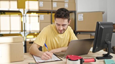 Young hispanic man ecommerce business worker using laptop writing on document at office