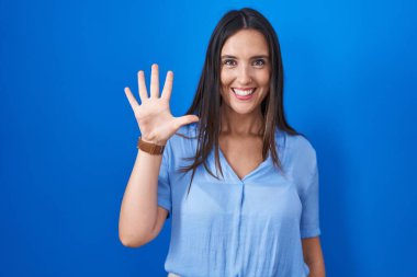 Mavi arka planda duran genç esmer kadın beş numarayı gösterip işaret ederken kendinden emin ve mutlu bir şekilde gülümsüyor.. 