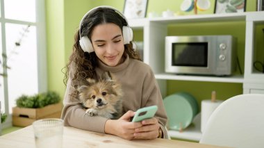 Yemek odasında akıllı telefon ve kulaklık kullanan köpekli genç İspanyol kadın.