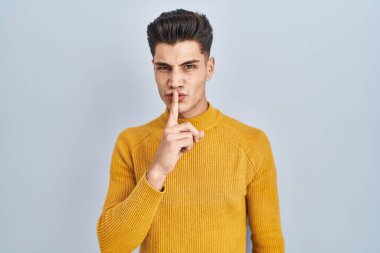 Mavi arka planda dikilen genç İspanyol adam parmakları dudaklarında sessiz olmak istiyor. sessizlik ve gizli konsept. 