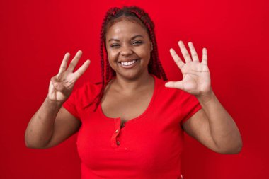 Kırmızı arka planda örülmüş saçları olan Afro-Amerikalı bir kadın sekiz numaralı parmaklarıyla gülümseyerek kendinden emin ve mutlu bir şekilde duruyor.. 