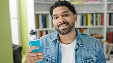 Afrikalı Amerikalı erkek öğrenci kütüphane üniversitesinde enerji dolu içecek şişesi tutuyor.