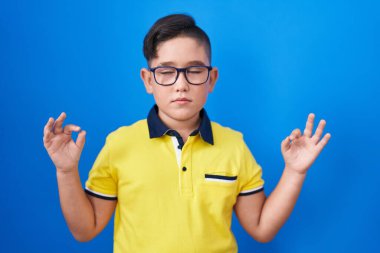 Mavi arka planda duran genç İspanyol çocuk rahatla ve gözleri kapalı gülümse parmaklarıyla meditasyon hareketi yapıyor. yoga konsepti. 