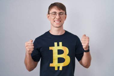 Başarı için heyecanlanan Bitcoin tişörtü giyen beyaz sarışın adam kollarını kaldırdı ve gözleri kapalı zaferi kutluyor. Kazanan konsept. 