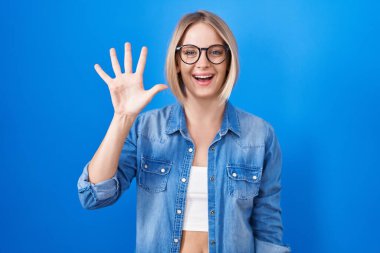 Young caucasian woman standing over blue background showing and pointing up with fingers number five while smiling confident and happy. 