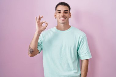 Pembe arka planda duran yakışıklı İspanyol adam gülümsüyor ve el ve parmaklarıyla 