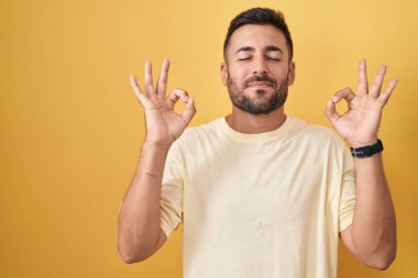 Sarı arka planda duran yakışıklı İspanyol adam rahatlamış ve parmaklarıyla meditasyon hareketi yaparken gözleri kapalı gülümsüyor. yoga konsepti. 