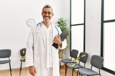 Orta yaşlı, gri saçlı, gülümseyen bir doktor. Klinik bekleme odasında kendine güvenen bir doktor.