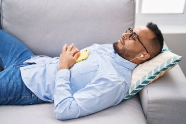 Genç İspanyol adam evdeki kanepede uzanmış müzik dinliyor.