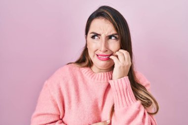 Young hispanic woman standing over pink background looking stressed and nervous with hands on mouth biting nails. anxiety problem. 