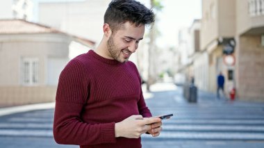 Genç İspanyol adam sokakta gülümseyen akıllı telefon kullanıyor.