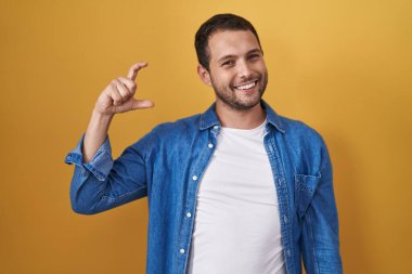 Hispanic man standing over yellow background smiling and confident gesturing with hand doing small size sign with fingers looking and the camera. measure concept. 