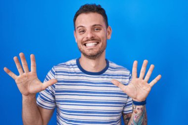 Young hispanic man standing over blue background showing and pointing up with fingers number ten while smiling confident and happy. 