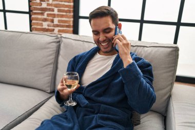 Young hispanic man talking on the smartphone and drinking wine at home