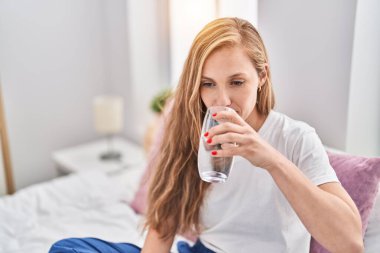 Young blonde woman drinking glass of water sitting on bed at bedroom