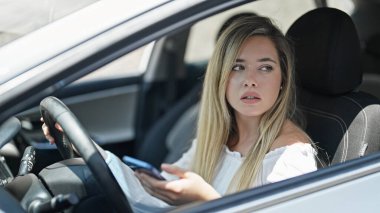 Genç sarışın kadın akıllı telefon kullanıyor. Sokakta arabada oturuyor.