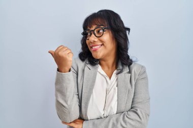 Hispanic woman standing over isolated background smiling with happy face looking and pointing to the side with thumb up. 