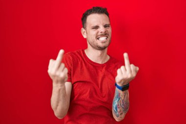 Young hispanic man standing over red background showing middle finger doing fuck you bad expression, provocation and rude attitude. screaming excited 
