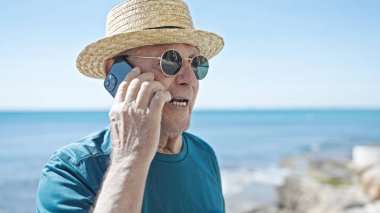 Kıdemli kır saçlı turist. Yazlık şapka takıyor. Deniz kenarında akıllı telefondan konuşuyor.