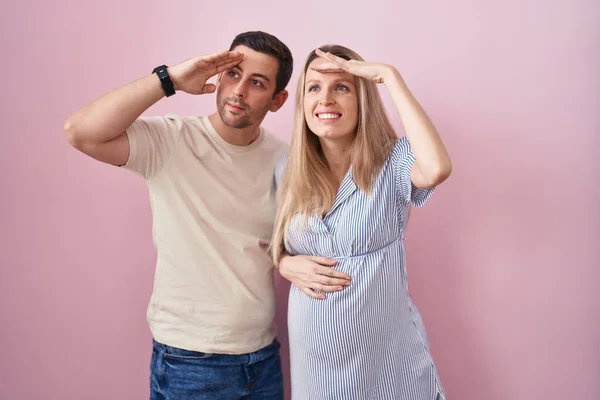 赤ん坊がピンクの背景の上に立っていることを期待している若いカップルは非常に満足し 頭の上に手で遠くを見て笑っています 検索の概念 — ストック写真