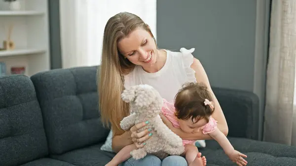 Mother and daughter sitting on sofa playing with teddy bear at home