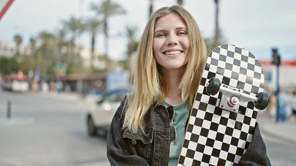 年轻的金发女子带着自信的笑容在街上举着溜冰鞋 — 图库照片
