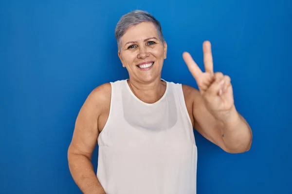 中年的高加索女人站在蓝色的背景上 微笑着看着摄像机 手指头在做胜利的标志 第二点 — 图库照片