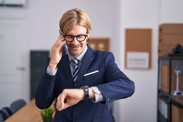 Young blond man business worker talking on smartphone looking watch at office