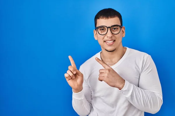年轻的阿拉伯男子 身穿随意的白色衬衫 戴着眼镜 面带微笑 看着摄像机 用双手和手指指向旁边 — 图库照片