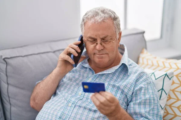 中年白髪の男話していますスマートフォン保持クレジットカード上のソファに家に座って — ストック写真