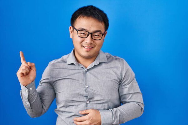 Young chinese man standing over blue background with a big smile on face, pointing with hand and finger to the side looking at the camera. 