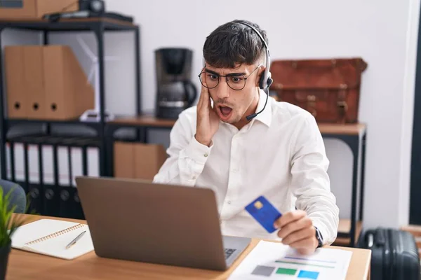 stock image Young hispanic man working using computer laptop holding credit card afraid and shocked, surprise and amazed expression with hands on face 