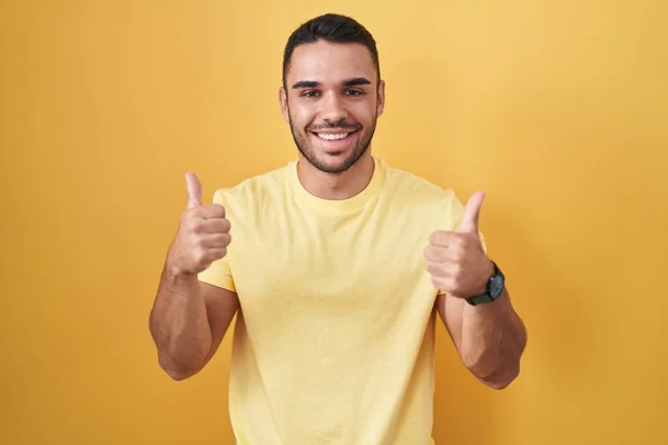 黄色の背景の成功のサインの上に立っている若いヒスパニック系の男は 手で積極的なジェスチャーを行うと 親指を笑顔と幸せ 陽気な表情と勝者のジェスチャー — ストック写真