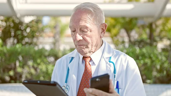 高级灰发男子医生在公园使用触摸板和智能手机 — 图库照片