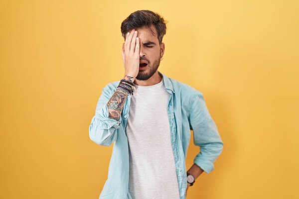 Young hispanic man with tattoos standing over yellow background yawning tired covering half face, eye and mouth with hand. face hurts in pain. 
