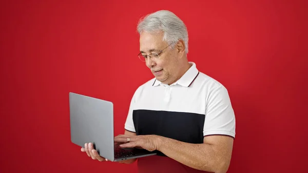 中年白发男子站在孤立的红色背景下使用笔记本电脑 — 图库照片