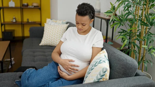 年轻的孕妇坐在沙发上 满脸愁容地摸着肚子 — 图库照片