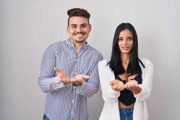 白い背景の上に立つ若いヒスパニック系のカップルが手を手のひらで一緒に受信またはジェスチャーを与える笑顔 保持し保護する — ストック写真