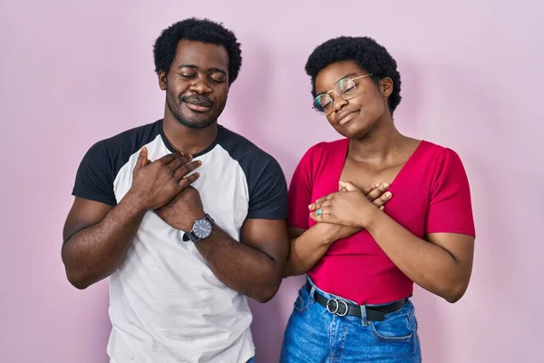 年轻的非洲裔美国夫妇站在粉红的背景上 双手放在胸前笑着 闭着眼睛 脸上挂着感激的手势 健康概念 — 图库照片